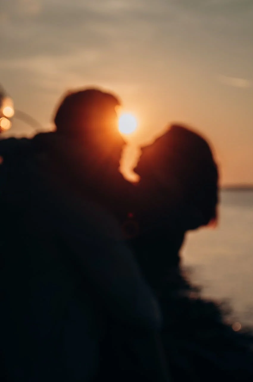 Siluetas desenfocadas de una pareja frente al sol del atardecer, junto al agua